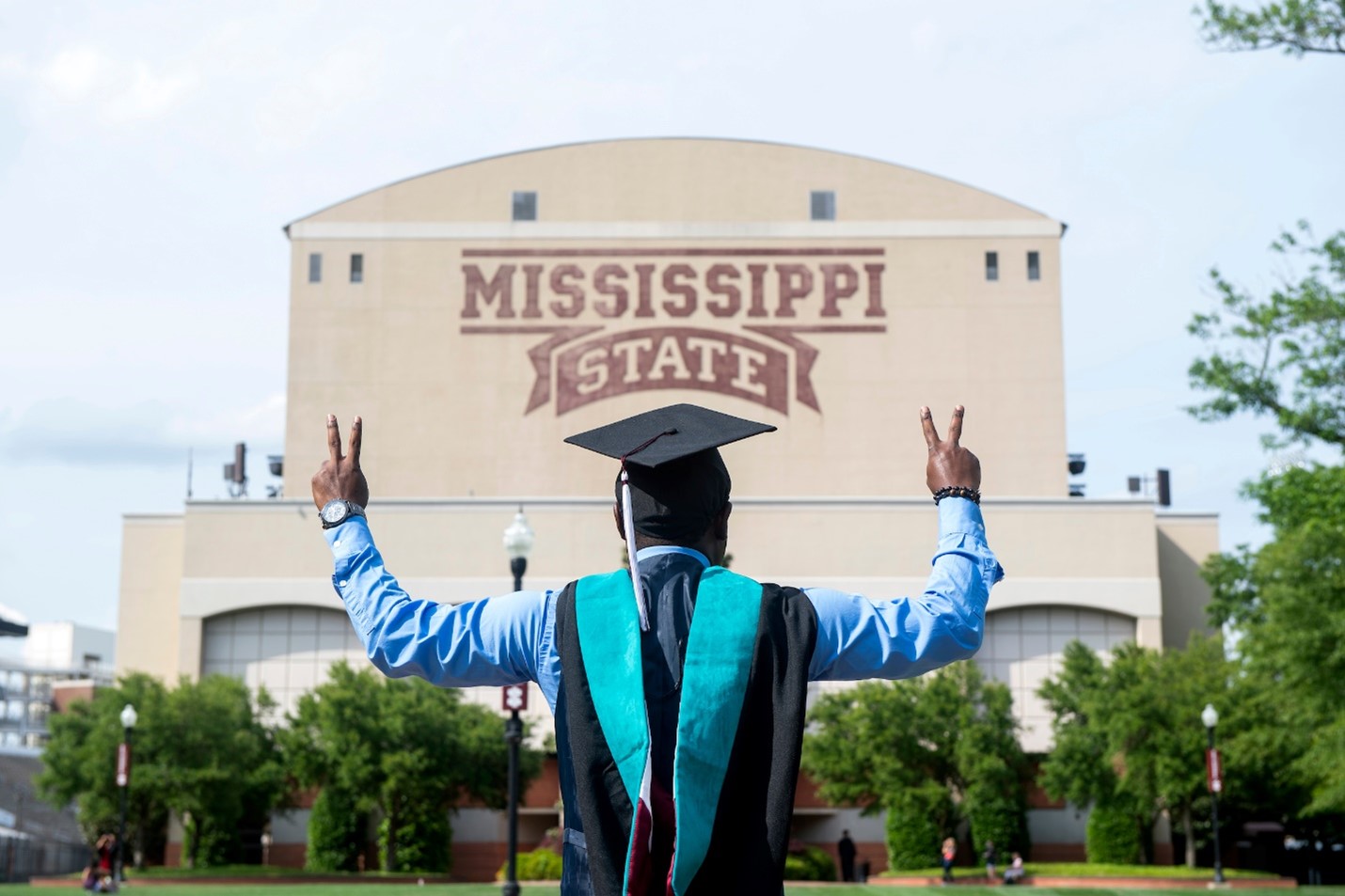 A student graduating from Mississippi State University stands in a graduation cap and gown outside Davis Wade Stadium