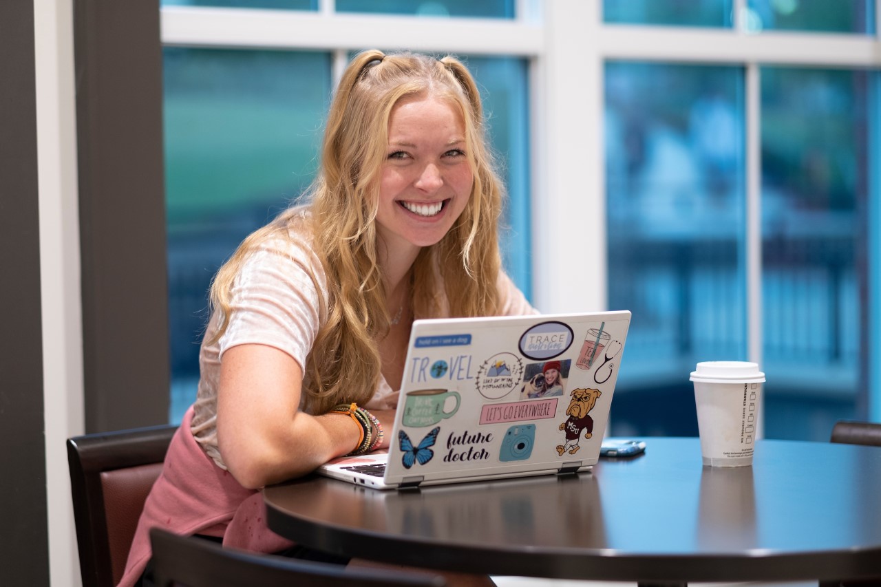 Smiling girl drinks coffee while using a laptop to study at Mississippi State University  