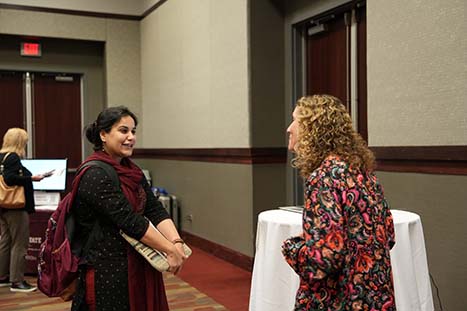 Two women are engaged in a conversation at an indoor event. The woman on the left is facing the camera, smiling, and dressed in traditional South Asian attire with a burgundy dupatta over a black top that has gold motifs. She carries a burgundy bag and holds what appears to be a brochure. The woman on the right, with her back to the camera, has curly hair and is wearing a colorful floral print dress. They appear to be standing near a conference or registration area, as indicated by a table with a white tablecloth in the background and another attendee behind them.